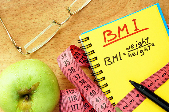 A notebook with "BMI" and the formula $\text{BMI} = \frac{\text{weight}}{\text{height}^2}$ written on a desk next to an apple, a pink measuring tape, glasses, and a black marker. Use this BMI Calculator to assess your health.