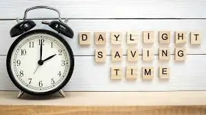 A black twin-bell alarm clock showing the time just after 1:00 is on the left, next to wooden tiles spelling out "DAYLIGHT SAVING TIME" on a white wooden background. The image relates to adjusting time, such as with a Time Calculator.